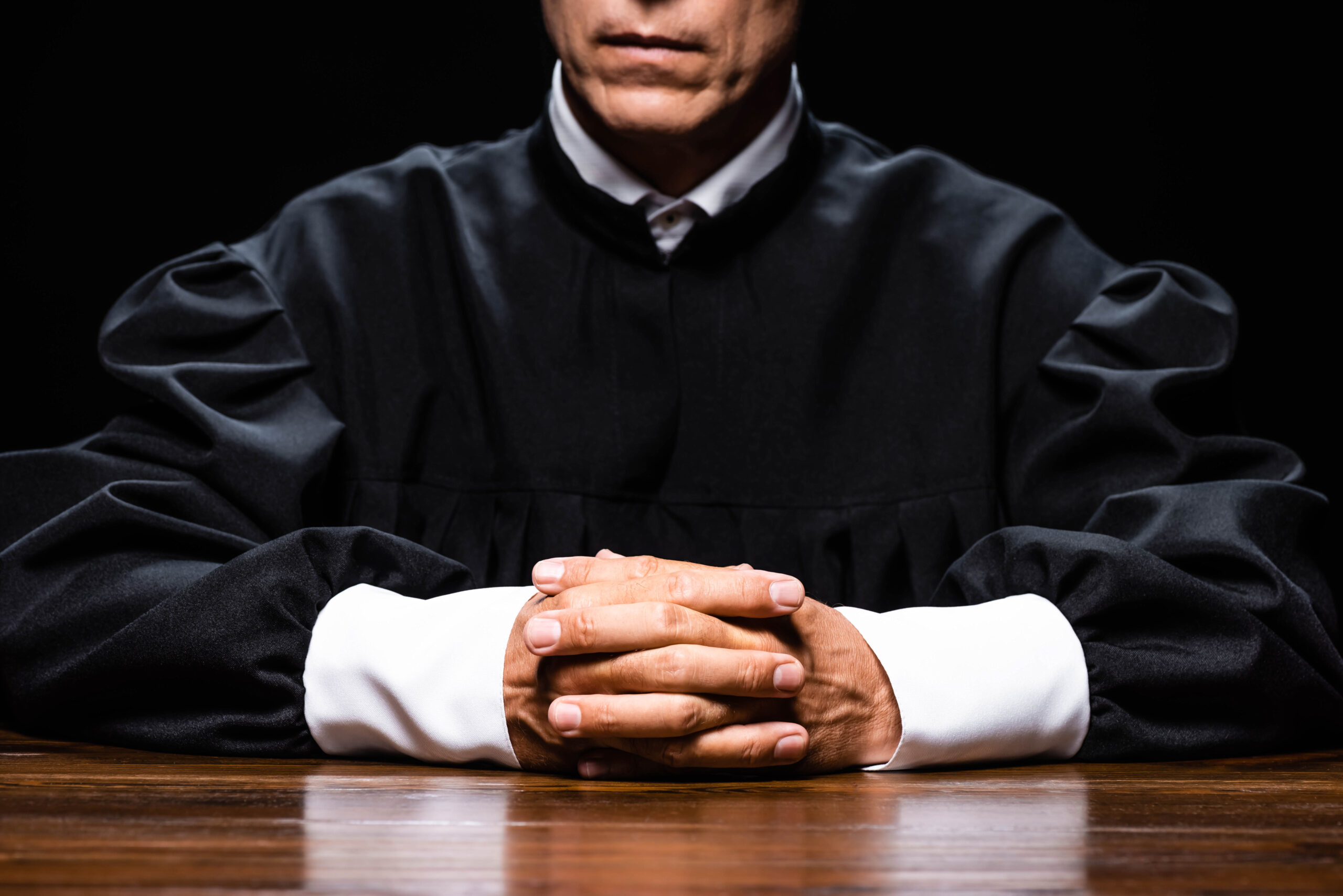 cropped view of judge in judicial robe sitting at table with clenched hands isolated on black